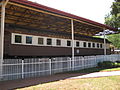 A brown train, viewed from the side, is at rest under a small open station. "1035 Rhodesia Railways" is written in gold letters over the train's windows. There is a white fence and a brick walkway in front of the train.