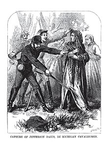man with shawl on head wearing an overcoat that looks almost like a dress being stopped by two soldiers.