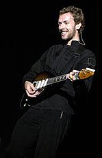Chris Martin wearing a black outfit and playing a brown electric guitar.