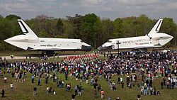 Enterprise and Discovery meet, rolling on their landing gear, seemingly nose to nose, along a paved path at Dulles airport. A throng of people standing on grass in the foreground has gathered to see the spectacle. Both orbiters have aeroshells covering the engine area.