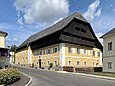 Ein zweistöckiges Haus mit Anbau und schwarzem Krüppelwalmdach. Das Haus ist gelb, die Fensterleibungen und die Ecksteine sind weiß.