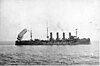 A dark gray, 3-masted ship with four funnels in a calm sea; a cigar-shaped balloon is tethered to her stern