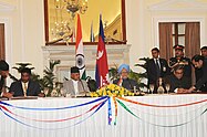 Mr. Sharat Singh Bhandari is shown at a formal table in front of Nepalese flags in his capacity as Minister of Tourism and Civil Aviation signing an air and railway infrastructure agreement with the Union Minister For External Affairs, Shri S.M. Krishna. A large mirror adorns the wall over an ornate mantelpiece in the background. An unknown person in a smart suit is also in attendance, standing in the right background of the photo.