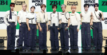 Nine men standing in a row on a stage dressed in coordinated outfits featuring white tops and dark blue pants with multiple straps and buckles. Some members are wearing accessories like gloves, arm bands, and headbands.