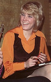 A young woman with blonde hair, wearing an orange blouse and a black vest