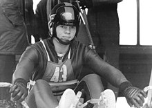A man wearing a skin-tight cold-protecting jumpsuit, gloves, and a full-face helmet with a lifted visor. He is sitted on a sled and there are people behind him.