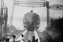A black and white photograph showing a ship's hull with no superstructure sliding down a slipway into a waterway