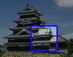 A two-storied tower connected on one side to a five-storied castle tower and on the other to a lower one-storied structure. All three structures have black wooden walls and are located on a platform of unhewn stones.