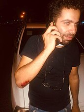 Mohammad Hossein Erfan on the phone, standing in front of a white car wearing a black shirt, a pair of glasses around his neck and visibly bleeding from his elbow.