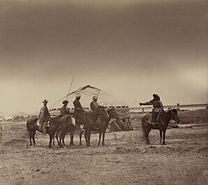 L'image montre une femme et 4 hommes à cheval face à une tente nomade. La femme tient un fouet et regarde en arrière. Cela est peut être un exemple de tradition de "capture" pour un mariage.