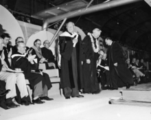 DeMille standing at a graduation ceremony in graduation robes among others sitting and applauding