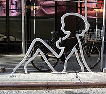 The outline of a silhouette of a naked woman leaning back, in galvanized steel, on a street curb. It has a bicycle with a shopping basket chained to it.