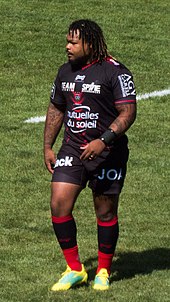 En pied, debout sur le terrain. Dreadlocks, moustache et barbe, tatouages sur les bras. Maillot noir, short noir, chaussettes rouge et noir.