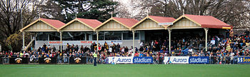 A grassed sporting oval with an old stand with a yellow and red roof, filled with spectators