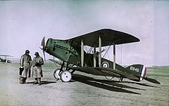Two men in long leather coats and leather helmets stand in front of a biplane in RAF colours.