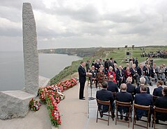 Ronald Reagan standing at a lectern and giving a speech to an audience who are seated in folding chairs in a u-shape around him. He stands at the center of the image in profile, facing his audience toward the right. Behind him, on the left side of the image, is the memorial at Pointe du Hoc, a rough stone obelisk. A floral arrangement is set on the ground in front of the monument.