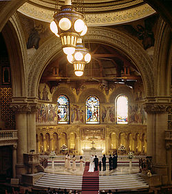 12 An interior view looking from high in the gallery, past two large arches which support the dome, and into the lofty semi-circular chancel. The building is of very large scale, and every part of the interior is covered with mosaic or carved decoration. In the chancel, a priest officiates for a bride and groom with eleven attendants.