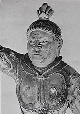 Head and shoulders portrait of a statue in front view. The right arm is raised, the hair sculpted with a top knot and the breast with armour. Narrow slit eyes and a facial expression as if frowning. Black and white picture.