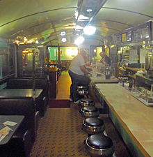 A long, narrow room with a counter and stools on the right at which two men are sitting and standing in the middle with a woman behind the counter a little further on. On the right are tables in booths. Above is a gently vaulted blue-green ceiling