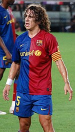 Curly haired man, looking tired, wearing a red and blue football shirt. In the background green grass is rarely visible