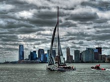 Bateau gris à la voile sous des nuages très sombres, devant des buildings. Dans le lointain, on distingue la statue de la Liberté.