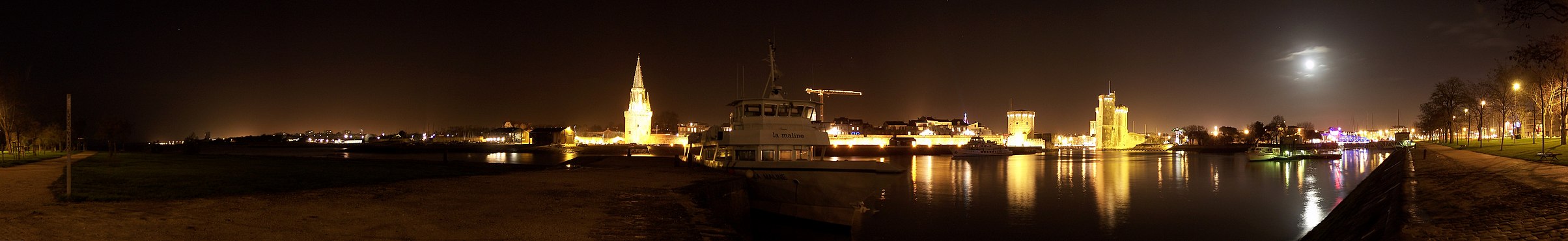 Panoramique nocturne des tours médiévales du Vieux-Port de La Rochelle depuis la digue du Bout-Blanc. La tour ayant un toit en forme de flèche est la tour de la Lanterne. Les deux autres tours, qui défendent l'entrée emblématique du Vieux-Port, sont la tour de la Chaîne (à gauche) et la tour Saint-Nicolas (à droite). Sous la lune, éclairé de lumière bleue, se trouve le pont levant du Gabut.