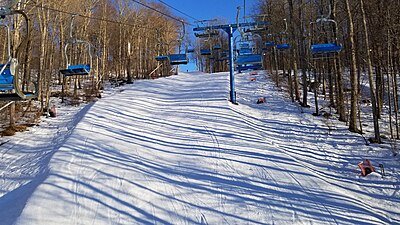 The image shows a two blue and gray ski double ski lifts sharing a tower on top of an uncrowded ski run covered in snow, with a blue ski