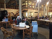 View of the reading room in the Maritime Research Center showing researchers, NPS uniformed staff as well as visitors, at tables consulting archives, books, and audio materials, Reference Librarian Gina Bardi at the Reference Desk in the background.