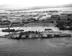 A ship on its side halfway underwater with cables strung to winches on the island behind it.