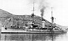 A gray, small battleship underway at slow speed in a harbor, a plume of smoke from its single stack visible against the sky and hills behind it