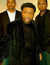 Andraé Crouch wearing black shirt and black jacket, and with a cap atop his head. He looks a little to the right, and stands with his mouth open. Behind him, two young Afro-American youths can be seen, with the one on right wearing spectacles.