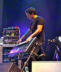 Derek Sherinian on stage playing a keyboard mounted on an angled keyboard stand. In the background are amplifiers, effects racks, a guitar rack and stage lighting mounted on the roof.