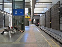 Underground grey tiled platform surrounded by concrete walls on either side. Light is coming down from above. There is a lift and set of stairs leading up to ground level.