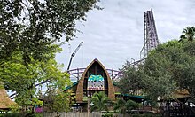 Iron Gwazi as pictured in July 2020 under construction, the façade of the main queue building from the entrance plaza can be seen. The lift hill is on the right with a construction crane on the left. Tree foliage surrounds the roller coaster in the foreground on the left and right.