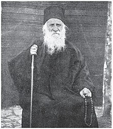 Gulabchev's father; he holds a stick and a necklace of beads. He is wearing traditional Orthodox Christian clothing.