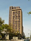 Ground-level view of a 20-story building with a dark brown facade; a setback is present near the roofline of the building. Dark windows are visible on the anterior side of the structure.