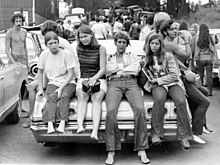 4 Woodstock festival attendees sitting on the trunk of a car facing the photographer. A crowd of people is walking down the road behind them.