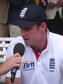 Headshot of man in white cricket gear and blue cap talking into a microphone.