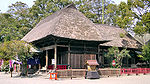 Three-quarter view of a wooden building with a thatched hip roof. Another structure of similar style extends from the back of the building.