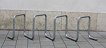 A free-standing bike rack, consisting of galvanized steel tubing bent into multiple square loops set at 45 degrees, on concrete slabs in front of a concrete wall
