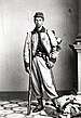 Full-body portrait of a white soldier with a rifle and bayonet by his side, standing on a crumpled flag. He is wearing baggy pants tucked into his boots, a short, decorative jacket buttoned at the top only, and a slouch cap.