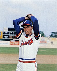 A man in a white baseball uniform with blue cap standing on a field with his hands together above his head posing in mid-pitching motion