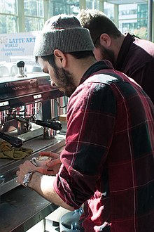 A man makes coffee in at a coffee shop. Sunlight streams through the windows.