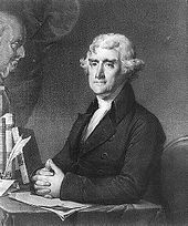 A man with tightly-curled gray hair wearing a black jacket and white shirt, sitting at a desk with hands folded on top of it