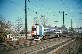 Stainless steel passenger rail cars with red and blue stripes horizontally across the windows. The visible end is covered with three vertical red, white, and blue stripes.