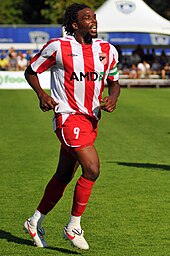 A black man with mid-length hair and a beard jogs across a grassy surface. He is wearing a red and white striped soccer jersey, red shorts and red socks.