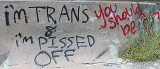 Graffiti on a concrete wall, in red and black. The black graffiti reads, "I'm trans & I'm pissed off". The red graffiti reads, "You should be".