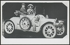blakc and white photograph of a young man and woman in an early 20th century open-top motor-car, with young child between them.