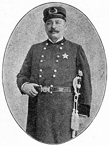 Black and white oval image of John T. Janssen in police uniform with tassels on the left side, grabbing onto his belt with his right hand.