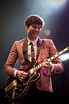 Mark Ronson performing on a stage wearing a pink suit and playing a black guitar
